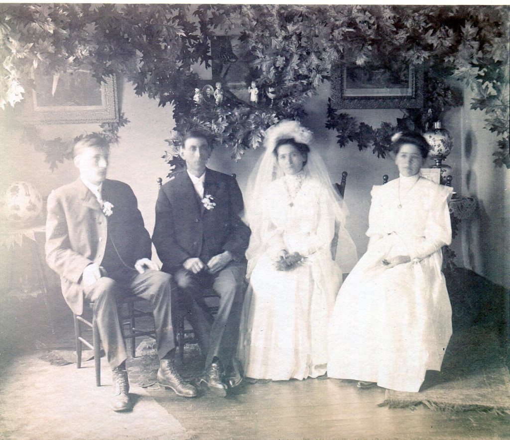 1906 Wedding Party consisting of Bride and Groom, Maid-of-Honor and Best Man in room decorated with leaves on the wall.